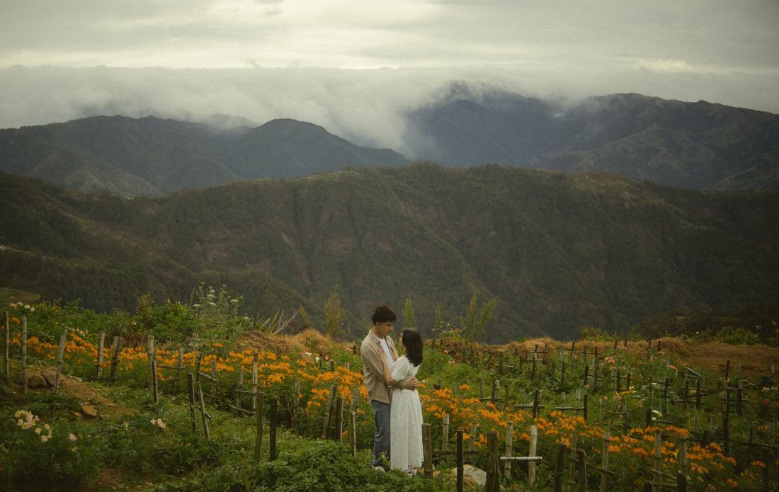Bryan and Irey holding each other in a field of marigolds, mountains folding away behind them.